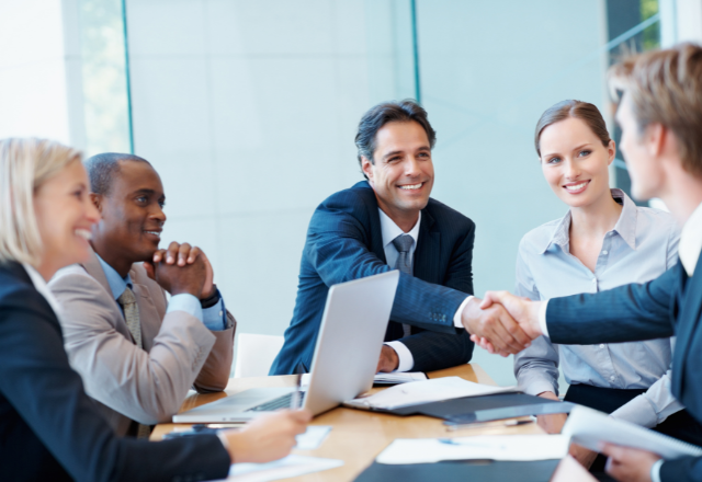 five people sitting at a table having a business meeting, two of the people are shaking hands